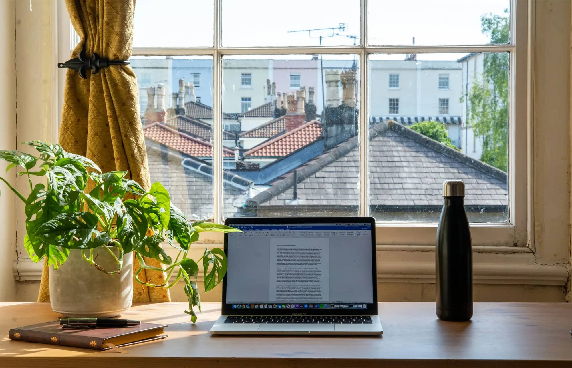 Table en bois près d’une grande fenêtre, ordinateur portable et plante verte dans la lumière du jour